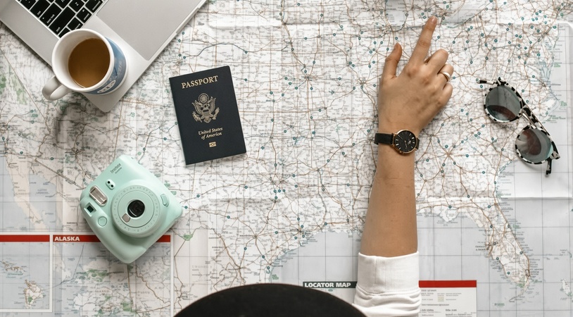 Top-down view of a large United States road map covered with travel items: an open laptop and white coffee mug in the top left, a U.S. passport and mint-green Instax camera near the center-left, a hand wearing a gold-and-black watch pointing at the eastern U.S., and patterned sunglasses on the right.