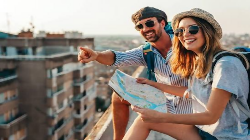 Smiling young couple on a city rooftop; man pointing into the distance while woman holds a map.