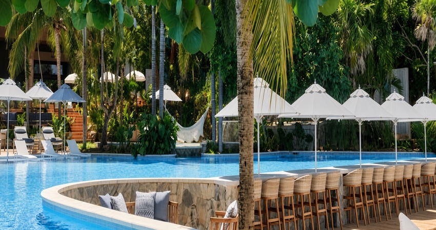 Luxury tropical resort swimming pool with turquoise water, wooden deck chairs under white umbrellas, and tall palm trees in the background.