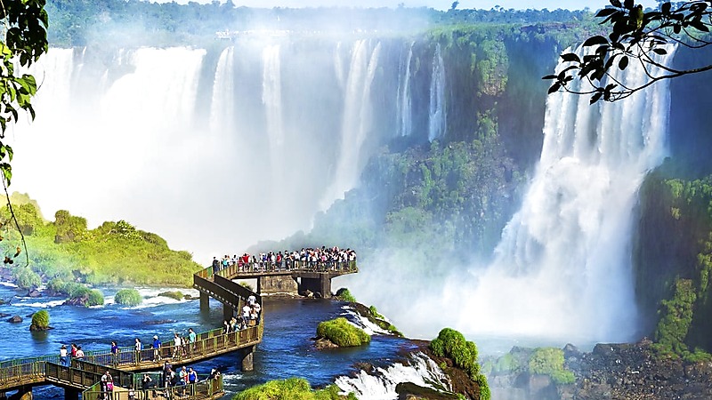 Tourists on walkways viewing the powerful cascading waterfalls of Iguazu Falls surrounded by lush green vegetation and mist.