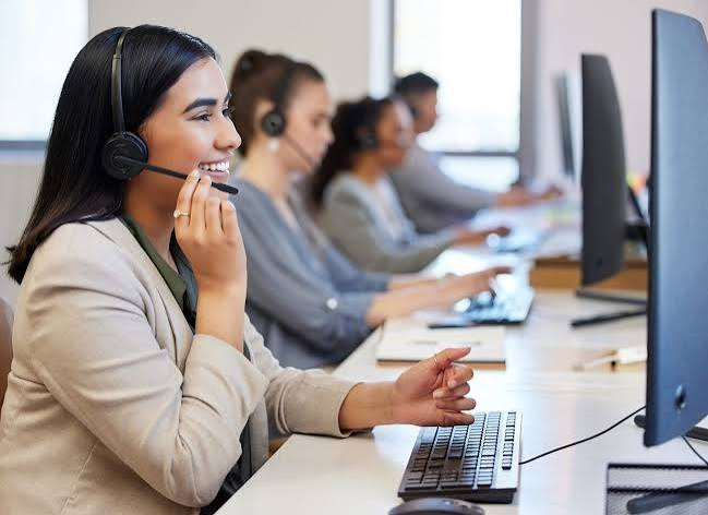 Smiling woman with headset working at a computer in a call center, colleagues in the background.